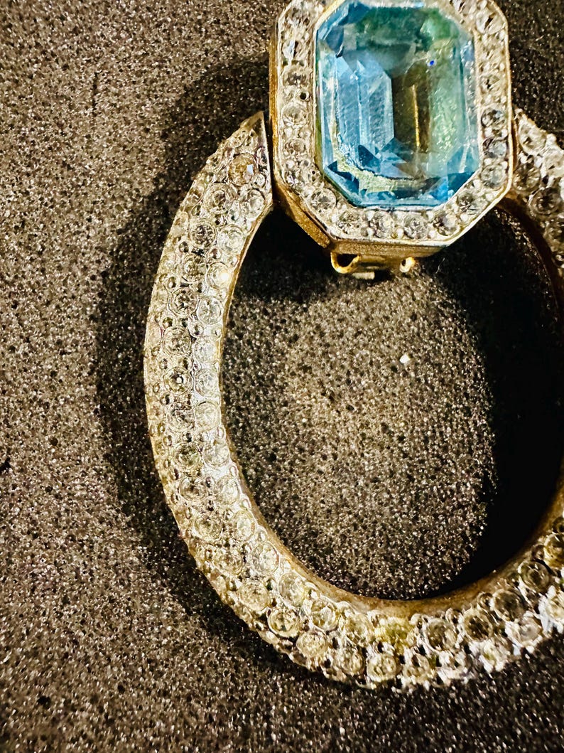 以下が含まれることがあります： 大きな青い宝石が、小さな透明な宝石で囲まれた金の指輪。指輪は、小さな透明な宝石の円形のバンドで飾られています。
