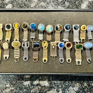 May include: A selection of vintage cufflinks on a dark grey tray. The cufflinks showcase diverse designs, including rectangular, square, and oval shapes, with gold, silver, and mesh straps. The cufflinks feature colourful gemstones and decorative elements.
