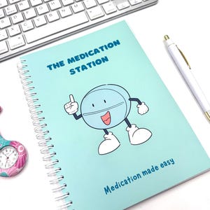 May include: A light blue spiral-bound notebook with the text "THE MEDICATION STATION" and a cartoon pill character. The notebook also has the text "Medication made easy". A white pen and keyboard are in the background.