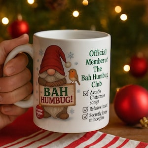 May include: White ceramic mug with a gnome illustration holding a sign that says "BAH HUMBUG!". The mug also features text that reads "Official Member of The Bah Humbug Club". A small bird is perched on the sign. Festive holiday theme.