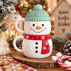 May include: A ceramic snowman mug set consisting of two stacked pieces. The top piece is a white mug with a light green knit-textured hat lid, a carrot nose, and a smiling face. The bottom piece is a white mug with a red scarf and black buttons.