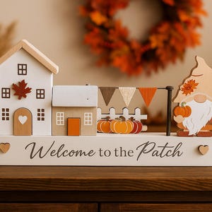 May include: A wooden autumn-themed decoration featuring miniature houses, pumpkins, and a gnome holding a pumpkin. The base reads "Welcome to the Patch". The color palette includes white, orange, and brown, evoking a fall harvest theme.