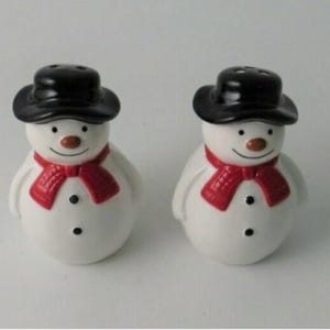 May include: Two snowman-shaped salt and pepper shakers. Each has a black hat, a red scarf, and a smiling face with a brown nose. The snowmen are white with black button details.