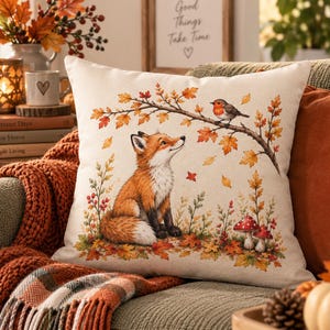 May include: A square throw pillow with a cream-colored background featuring a printed illustration of a red fox sitting in a forest setting with autumn leaves, a small bird perched on a branch, and two red and white spotted mushrooms at the base.