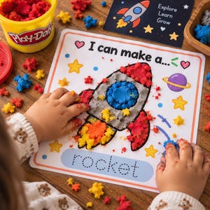 Könnte beinhalten: Eine quadratische Aktivitätsmatte mit dem Umriss einer Rakete, die mit bunter Knete gefüllt werden kann. Die Matte enthält den Text I can make a... und rocket in gepunkteter Schrift, umgeben von Sternen und einem Planeten. Ein gelber Play-Doh-Becher ist sichtbar.
