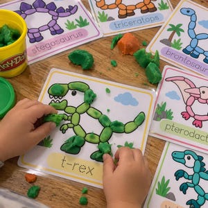 Könnte beinhalten: Ein Set laminierter Dinosaurier-Aktivitätskarten mit Umrissen von Stegosaurus, Triceratops, Brontosaurus, Pterodactylus, Velociraptor und T-Rex, wobei grüne Knete den T-Rex ausfüllt und ein gelber Play-Doh-Becher daneben steht.