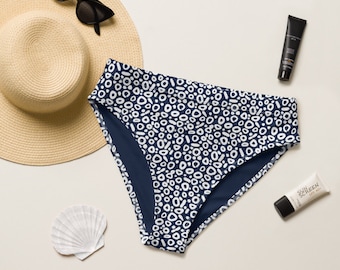 May include: Navy blue bikini bottoms with a white abstract pattern. The bottoms are on a white surface with a straw hat, seashell, and sunscreen.