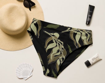 May include: Black and green bikini bottom with a leafy print. The bikini bottom is on a white surface with a straw hat and sunscreen.