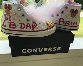 May include: A pair of white high-top sneakers featuring gold glitter mouse ear silhouettes, pink lettering reading 2ND B-DAY and ARIA, gold stud embellishments on the toe caps, and decorative light pink tulle bows attached to the laces.