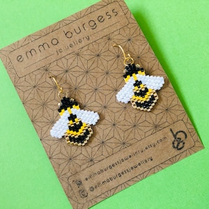 Pode incluir: Brincos de abelha feitos à mão com ganchos dourados. Os brincos são feitos de pequenas contas pretas, brancas e amarelas, dispostas para formar uma abelha. São exibidos em um cartão marrom com o texto "emma burgess jewellery".