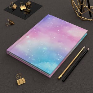 May include: A hardback journal featuring a galaxy design in blue, purple, and pink, with white star details. Gold and black pencils and gold binder clips are placed nearby.