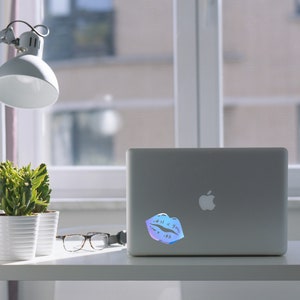 May include: A silver laptop computer with a holographic sticker of a pair of lips on the lid. The laptop is sitting on a white desk with a potted succulent plant and a pair of glasses.