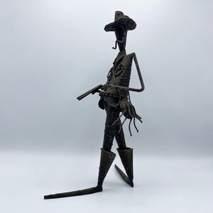 May include: A rustic metal sculpture of a cowboy figure, complete with a hat, boots, and a gun. The sculpture has a textured, weathered appearance, suggesting it is handmade. The cowboy is depicted in a walking pose, adding a dynamic element to the artwork.