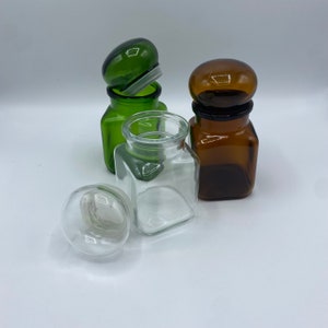 May include: Three glass jars with lids. One jar is green, one is brown, and one is clear. The jars are all square shaped and have a rounded lid.