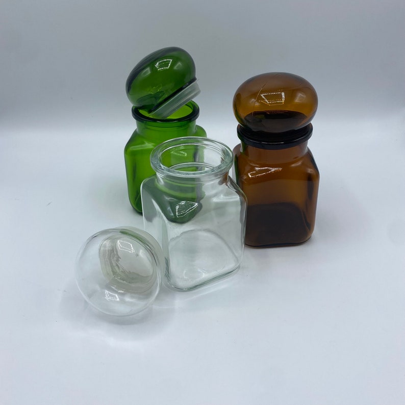 May include: Three glass jars with lids. One jar is green, one is brown, and one is clear. The jars are all square shaped and have a rounded lid.