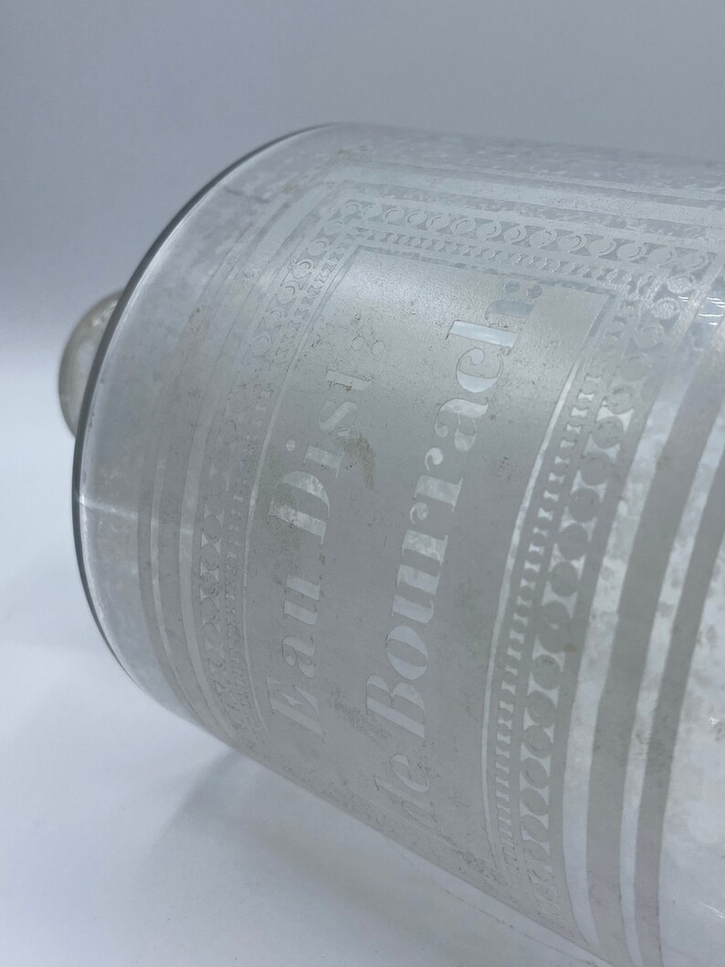May include: A clear glass bottle with an etched design. The bottle is labeled "Paul Dis. de Bourjac".