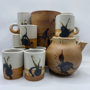 Może przedstawiać: Kolekcja ręcznie robionej ceramiki stołowej. Zestaw zawiera kubki, szklanki i czajniczek, wszystkie w neutralnej palecie kolorów bieli i brązu. Każdy element posiada unikalny czarny wzór rozprysków.