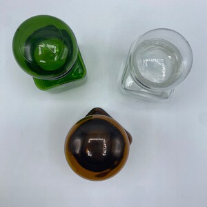 May include: Three glass bottle stoppers with different colored glass tops. The stoppers are in the shape of a sphere and are sitting on a white surface.