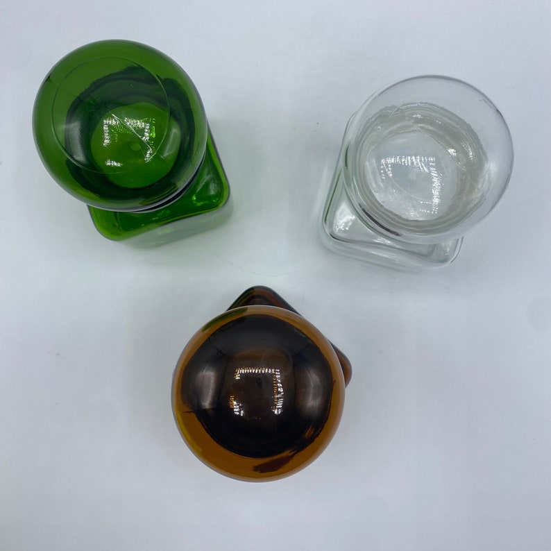 May include: Three glass bottle stoppers with different colored glass tops. The stoppers are in the shape of a sphere and are sitting on a white surface.