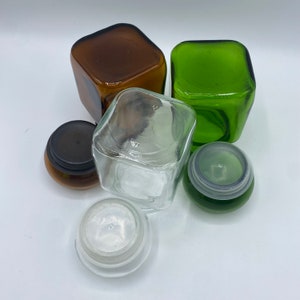 May include: A set of four glass jars with lids. Two jars are green, one is brown, and one is clear. The jars are square shaped and have a vintage look.