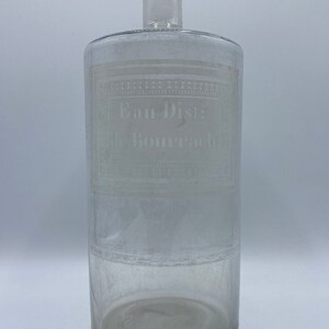 May include: A clear glass bottle with a cylindrical body and a narrow neck. The bottle has an etched label that reads "Eau Dist: de Bourrache".