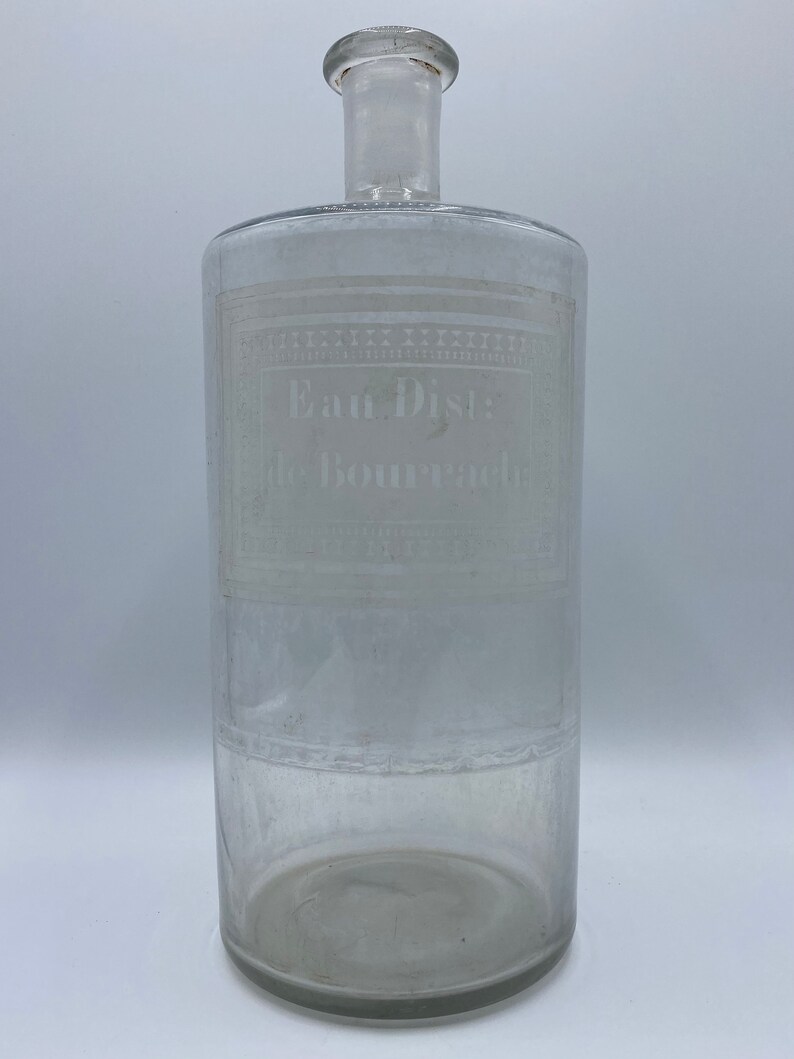 May include: A clear glass bottle with a cylindrical body and a narrow neck. The bottle has an etched label that reads "Eau Dist: de Bourrache".