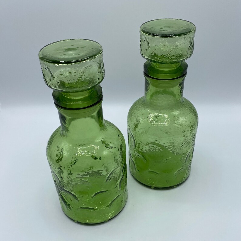 May include: Two green glass bottles with textured surfaces and stoppers. The bottles are empty and have a vintage look.