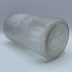 May include: A clear glass bottle with a white label. The label has a design of a square with a circle inside. The bottle is lying on its side.