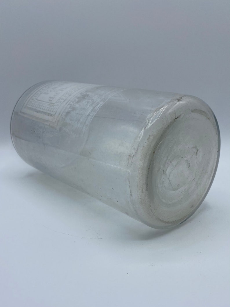 May include: A clear glass bottle with a white label. The label has a design of a square with a circle inside. The bottle is lying on its side.