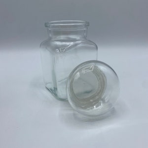 May include: A clear glass jar with a square body and a round lid. The jar is empty and the lid is off.