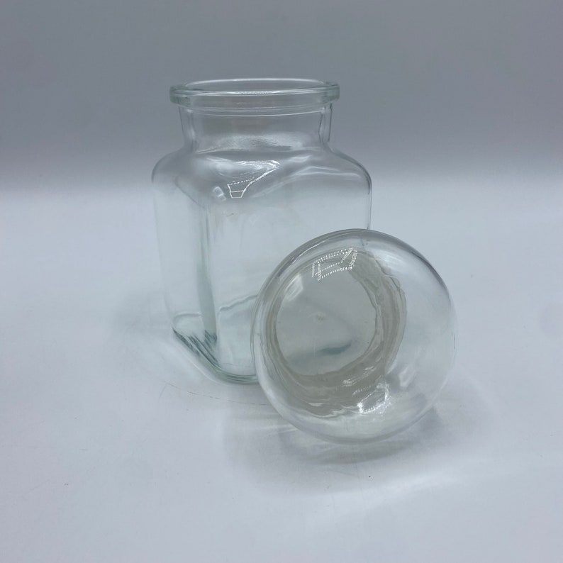 May include: A clear glass jar with a square body and a round lid. The jar is empty and the lid is off.