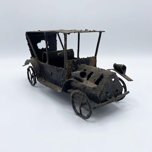 May include: A miniature, antique-style car sculpture. The model is made of dark metal with a rusted appearance, featuring a detailed frame, wheels, and a roof. The car has a vintage aesthetic, with visible cutouts and a weathered finish.