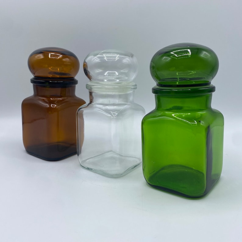 May include: Three glass jars with lids. The jars are square shaped and have different colored glass. One jar is brown, one is clear, and one is green.