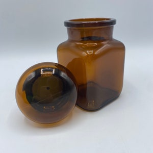 May include: A brown glass jar with a round lid. The jar is square and has a wide mouth. The lid is made of glass and has a smooth surface.