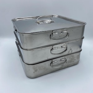 Op de afbeelding: Stapel van drie rechthoekige roestvrijstalen containers. Elke container heeft een handvat. De bovenste container heeft een handvat op het deksel. De containers zijn gemaakt van roestvrij staal.