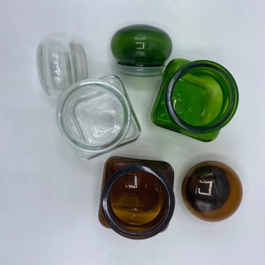 May include: Four glass jars with lids. Two jars are clear glass, one is green glass, and one is brown glass. The jars are all different sizes and shapes.