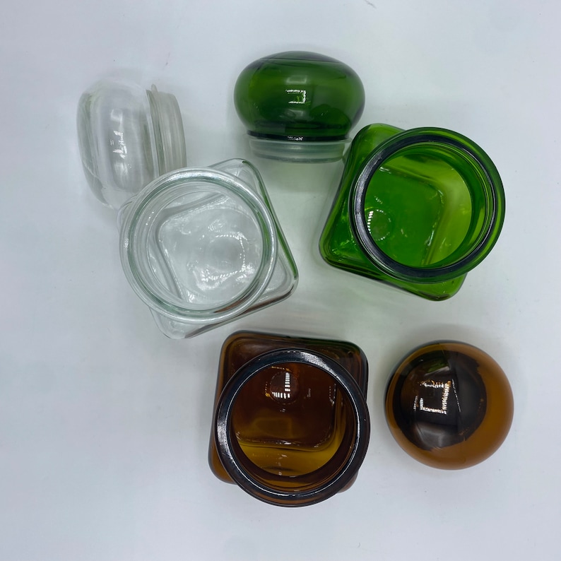 May include: Four glass jars with lids. Two jars are clear glass, one is green glass, and one is brown glass. The jars are all different sizes and shapes.