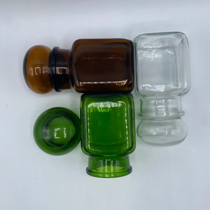 May include: Four glass jars with lids. Two jars are green, one is brown, and one is clear. The jars are arranged in a square shape.