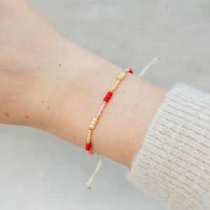 Könnte beinhalten: Ein zartes Armband mit kleinen roten, rosa und cremefarbenen Perlen, die auf einer weißen Schnur aufgefädelt sind.
