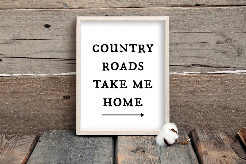 May include: A framed print with the text "COUNTRY ROADS TAKE ME HOME" and an arrow pointing to the right. The print is on a wooden surface.