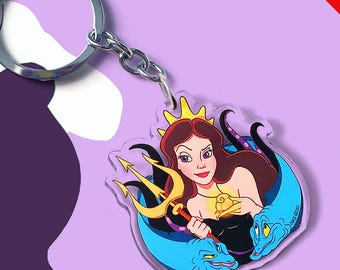 May include: An acrylic keychain featuring a cartoon illustration of a character with long brown hair, a gold crown, and a gold trident, flanked by two blue eel-like creatures. The character wears a black top and a gold shell necklace.