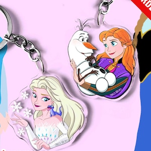 Op de afbeelding: Twee acryl sleutelhangers met cartoonillustraties. Eén sleutelhanger toont een blond personage met een kleine blauwe hagedis en witte sneeuwvlokken. De andere toont een roodharig personage dat een witte sneeuwpop met een oranje neus vasthoudt.