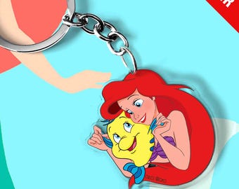 May include: A circular acrylic keychain featuring a colorful illustration of a red-haired mermaid character embracing a small yellow and blue fish. The keychain is attached to a silver-toned metal split ring by a short chain link connector.