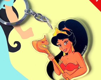 May include: A clear acrylic keychain featuring a cartoon illustration of a character with dark hair, a gold crown, and a red top, holding a golden cobra-headed staff. The keychain includes a silver metal ring and a short chain attachment.