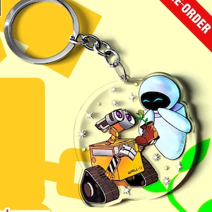 Puede incluir: Un llavero de acrílico transparente con una ilustración colorida del robot WALL-E sosteniendo una bota marrón con una pequeña flor amarilla, junto al robot EVE. El diseño incluye detalles de estrellas brillantes y una anilla de metal plateado.