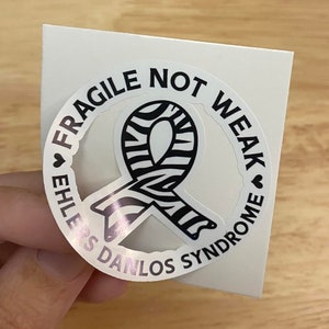 May include: A white sticker with a black and white zebra striped ribbon in the center. The sticker is round and has the text "Fragile Not Weak Ehlers Danlos Syndrome" around the ribbon.