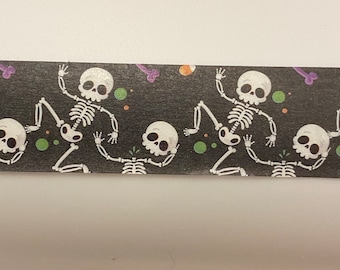 May include: A black and white patterned washi tape with a repeating design of skeletons dancing. The skeletons are white with black eyes and are on a black background with green and purple accents.