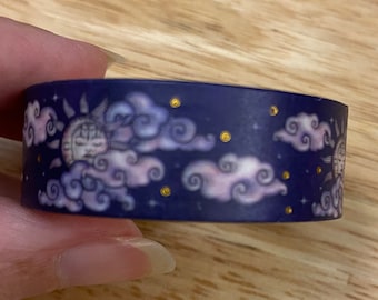 May include: A roll of washi tape with a dark blue background featuring a repeating pattern of white clouds, gold stars, and a sun and moon design.