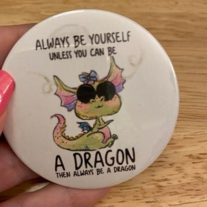 May include: White round button with a green dragon with pink wings and a blue bow. The text on the button reads "Always be yourself unless you can be a dragon then always be a dragon."