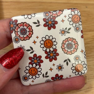 May include: A white square-shaped compact mirror with a colorful floral pattern. The pattern includes red, orange, and yellow flowers with peace signs and smiley faces.